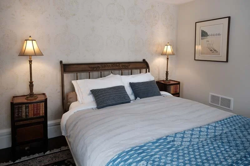 A neatly made bed with white and gray bedding, two dark gray pillows, flanked by matching wooden-nightstands with table lamps, framed artwork on the wall, and a vent near the floor.