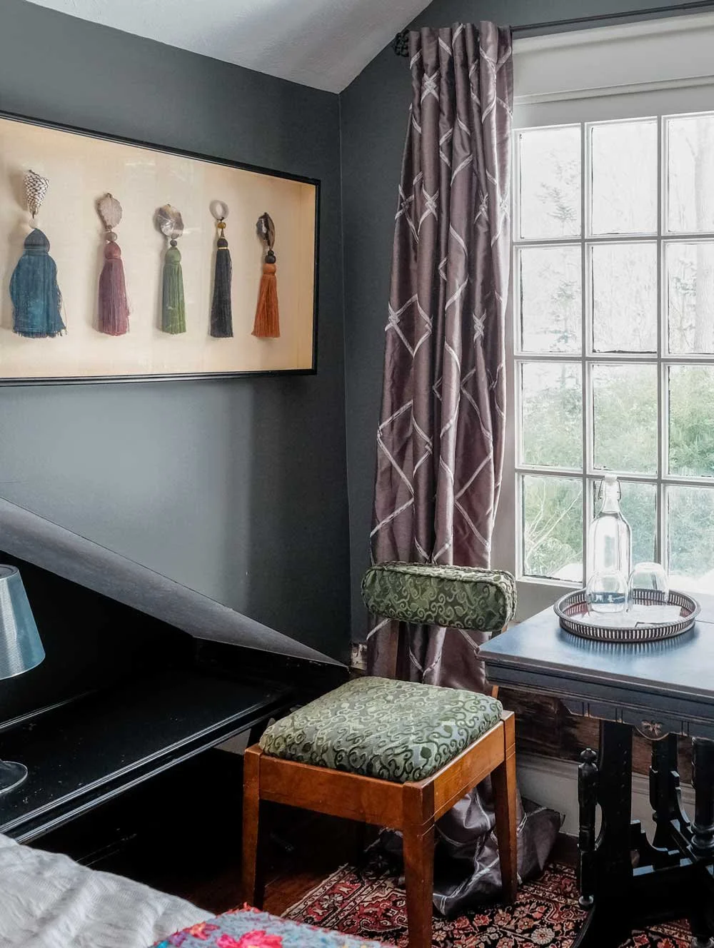 Corner of a room with a window and curtain, framed art with tassels, a piano, a wooden chair with patterned green upholstery, and a side table with a tray holding glass bottles.