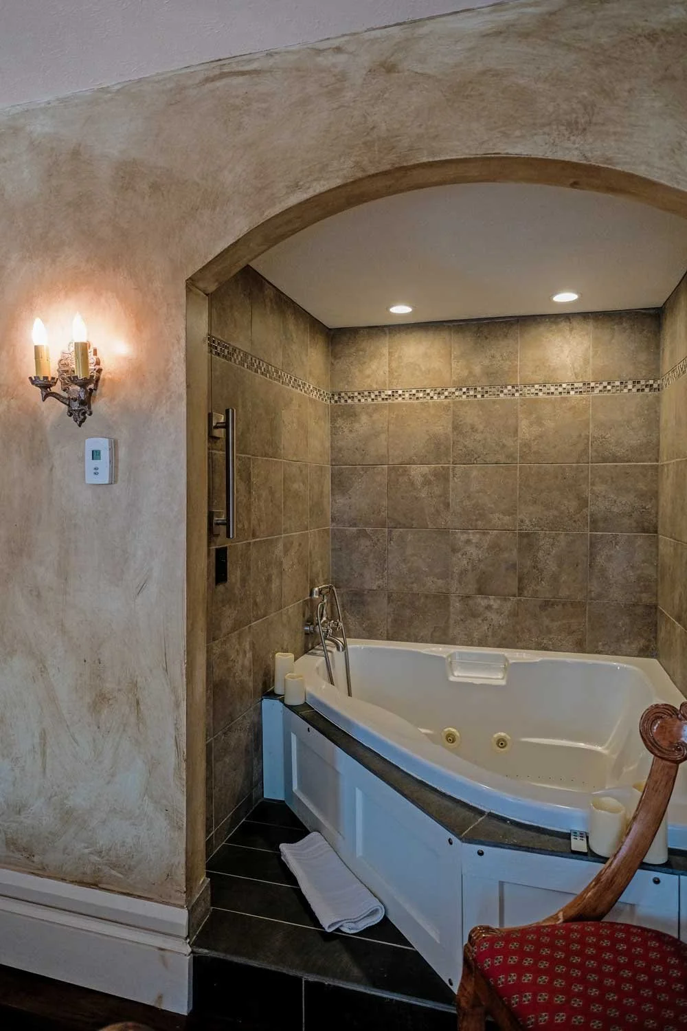 Bathroom with a corner whirlpool bathtub, beige and brown tiles, candles, a towel on the floor, a wooden chair with a red cushion, and wall-mounted lighting.