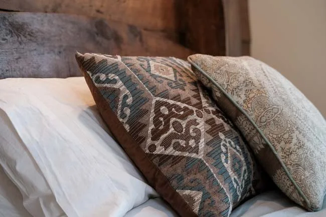 Close-up of a bed with white sheets and two decorative pillows, one with a dark patterned fabric and the other with a light patterned fabric, against a wooden headboard.