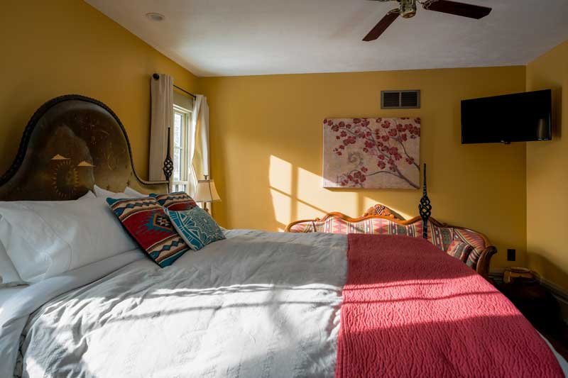 Sunlight shining into a yellow-walled bedroom with a large bed, decorative pillows, a side table with a lamp, a window, a wall art piece, a flat-screen TV, and a ceiling fan.