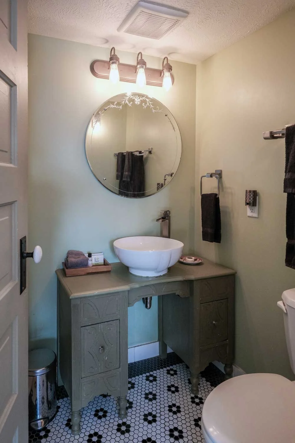 Bathroom with a round mirror, a white vessel sink on a green vanity, and black towels. The floor has black and white hexagonal tiles with floral patterns. There is a light fixture with three bulbs above the mirror and a small trash can beside the vanity.