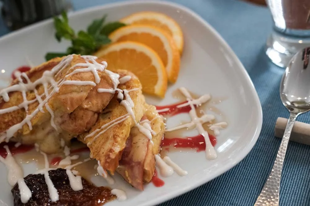 Plate of fried chicken drizzled with white sauce, garnished with orange slices and greenery, with a glass of beverage and a spoon on the side