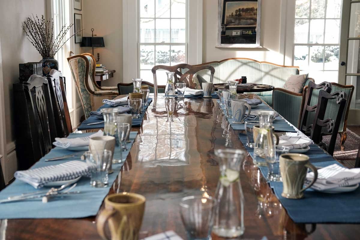 A dining room with a large wooden table set for a meal, with tableware, glasses, and napkins, surrounded by chairs and a vintage sofa, with large windows letting in natural light.
