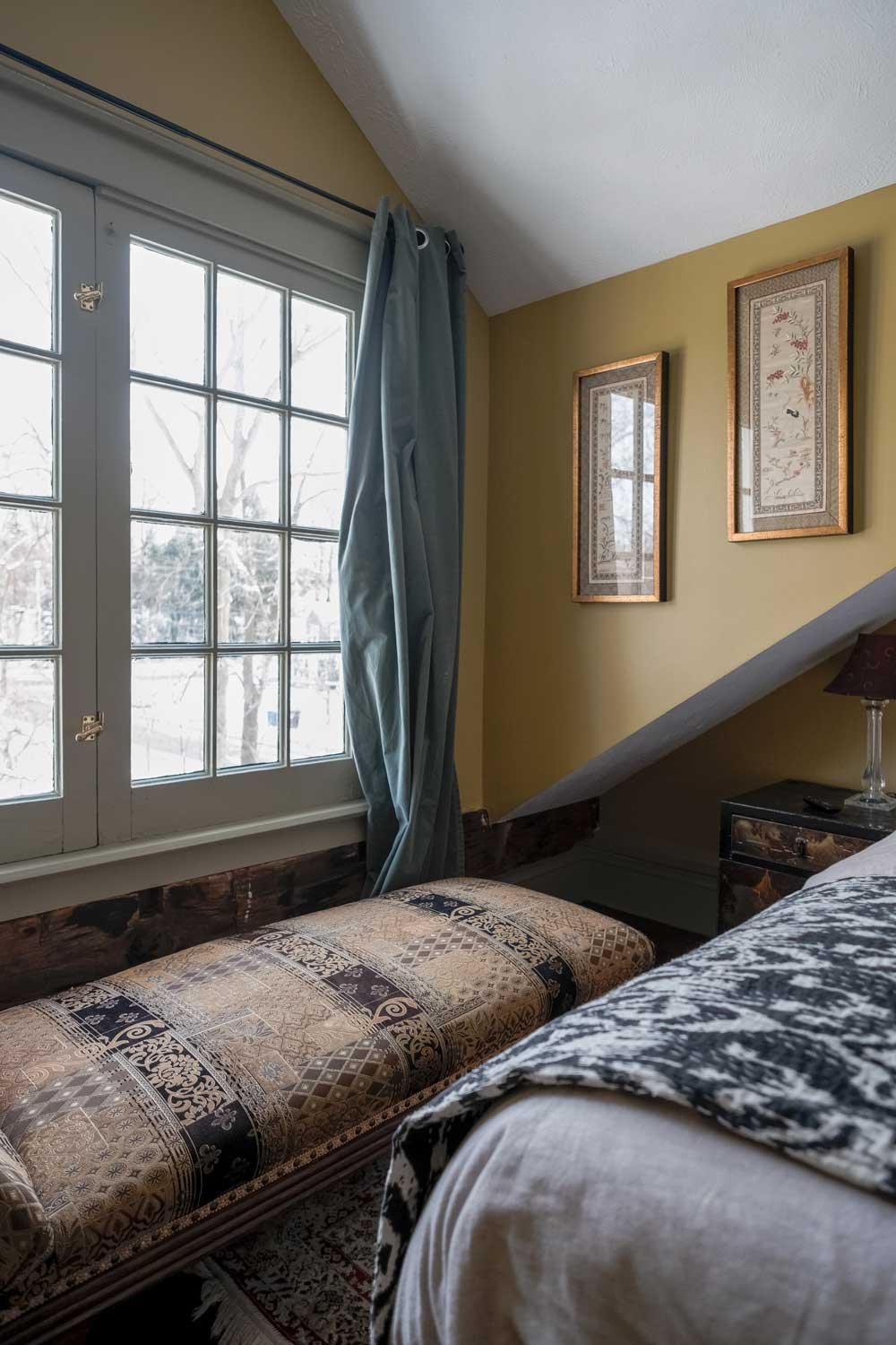 A cozy bedroom corner with a large window with grid panes, blue curtains, framed botanical art on yellow walls, a patterned bench under the window, and part of a bed with a black and white patterned blanket and a wooden nightstand with a glass lamp.