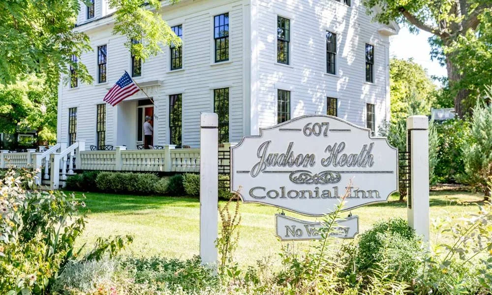 White colonial inn building with an American flag, surrounded by green trees and grass, with a sign reading "Judson Heath Colonial Inn" and "No V" in the foreground.