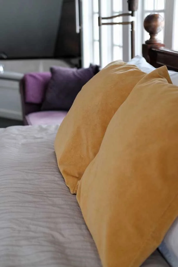 Close-up of two yellow pillows on a neatly made bed with a gray and purple couch in the background, near a window with white blinds.