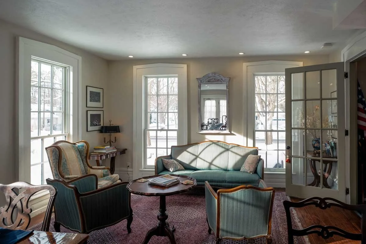 Living room with large windows, vintage furniture including green and patterned armchairs, a sofa, a round wooden table with magazines, and a door opening to another room, with snow outside visible through the windows.