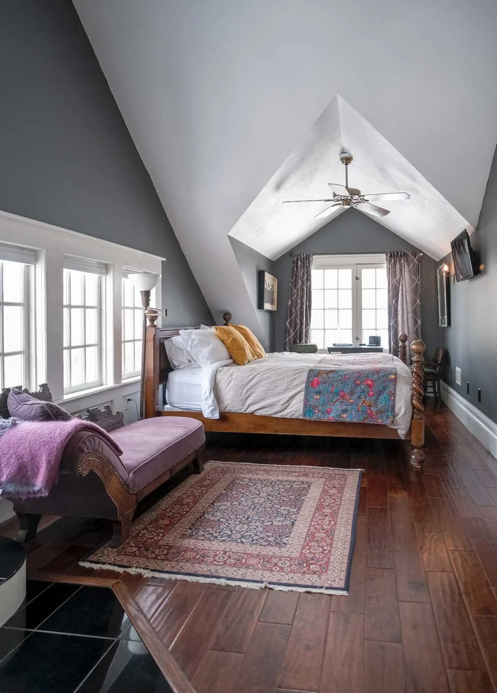 A cozy bedroom with a sloped ceiling painted in dark gray, featuring white trim, a wooden bed with white bedding and yellow pillows, a purple chaise lounge with a pink throw, a traditional rug, and windows with purple curtains, illuminated by natural light.