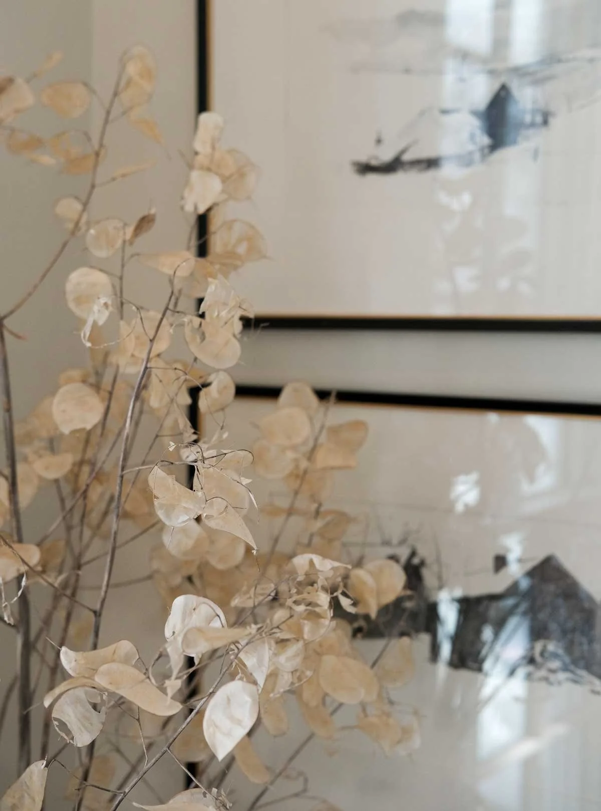 Close-up of dried, beige-colored leaves and branches in front of two framed abstract art pieces on a wall.
