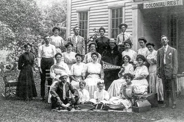 Group of young people and two adults posed outside a building with a sign that reads 'Colonial Rest', holding a sign that says 'Saugatuck.'