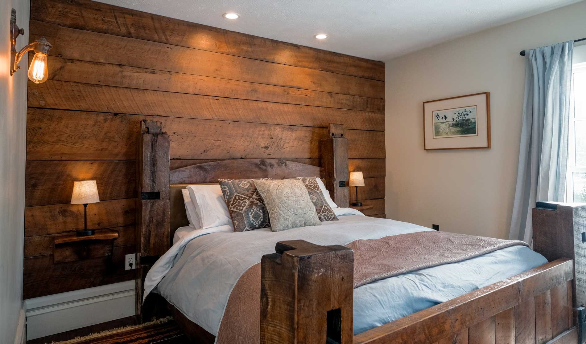 A cozy bedroom featuring a large wooden bed with a high headboard and a rustic wooden accent wall behind it. The bed has white linens, decorative pillows, and a pinkish bed runner. There are two small bedside lamps on either side, a framed artwork on the wall, and a window with light-colored curtains letting in natural light.