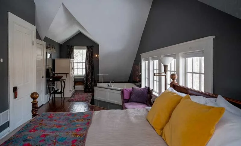A cozy attic bedroom with sloped ceilings painted dark gray, featuring a bed with colorful pillows, a pink armchair, a small white bathtub, a window with curtains, and a desk area.