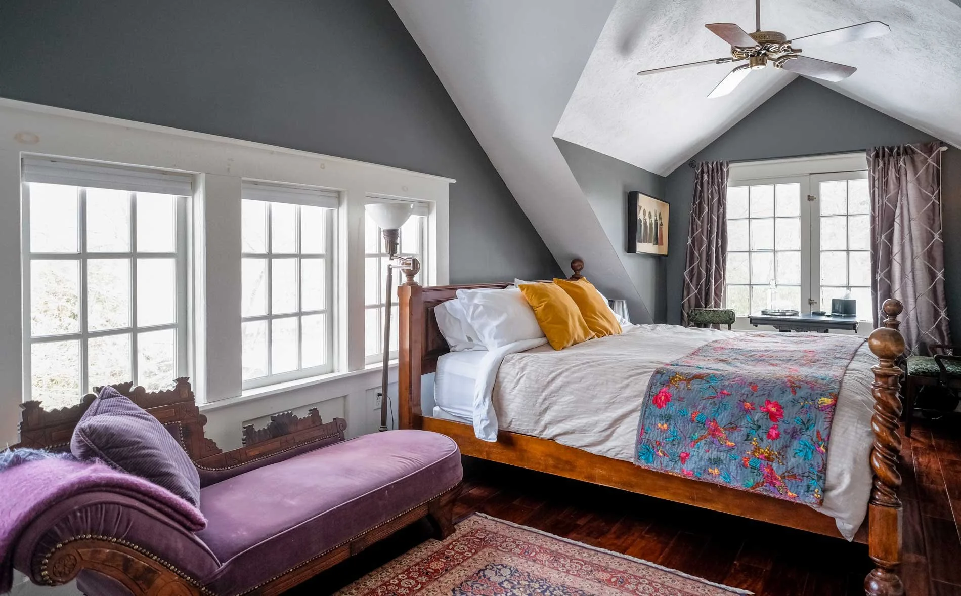 Bedroom with a wooden four-poster bed, white bedding, yellow pillows, colorful quilt, purple chaise lounge, windows with white frames, patterned curtains, a ceiling fan, and framed artwork.