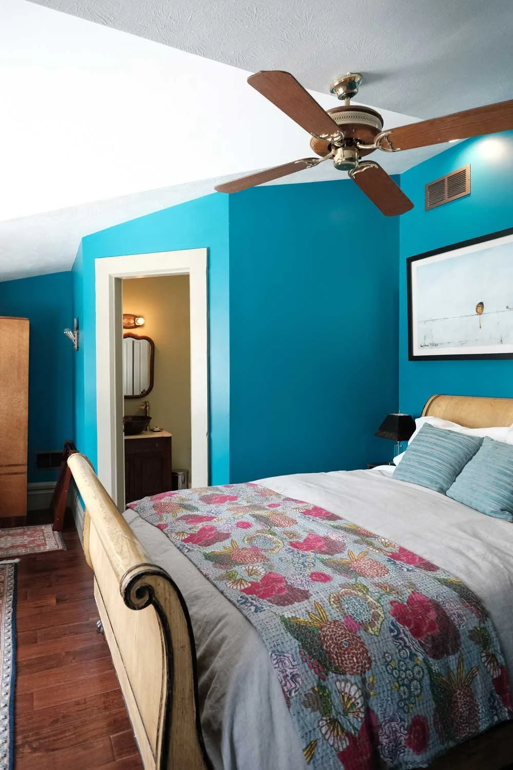 A bedroom with a wooden bedframe, white bedding, and blue pillows, featuring a colorful quilt. The room has a vibrant blue wall, a framed art piece, a black lamp, and a ceiling fan with wooden blades. There is a doorway leading to a bathroom with a sink, mirror, and light. The floor is hardwood, with a small rug near the bed.