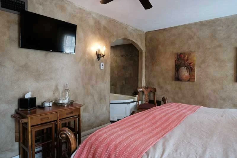 A bedroom with a flat-screen TV mounted on a textured beige wall, a wooden desk with a phone, water pitcher, and tissue box, a wall-mounted light fixture, a bathtub visible through an arched doorway, a chair, and a bed with a pink-striped throw blanket.