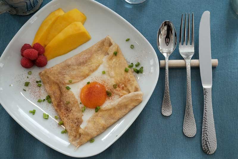 Breakfast plate with fried egg, crepe, pineapple slices, and raspberries, set on a blue table with utensils.