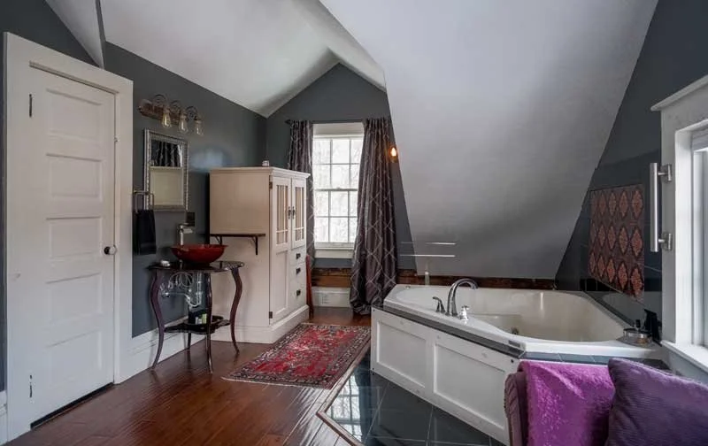 A cozy attic bathroom with dark blue walls, wooden flooring, a bathtub built into a white surround, a purple towel on a purple chair, a window with curtains, a white cabinet, a sink with a red basin, and a decorative mirror