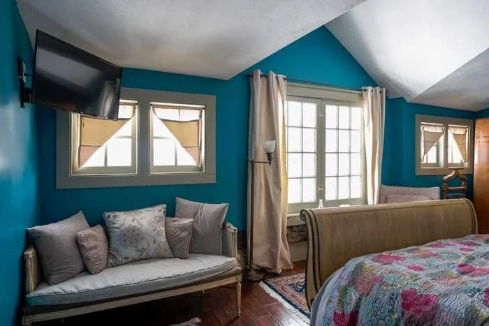 A cozy bedroom with blue walls, a bed with floral bedding, a vintage sofa with pillows, a wall-mounted TV, and large windows with white curtains letting in natural light.