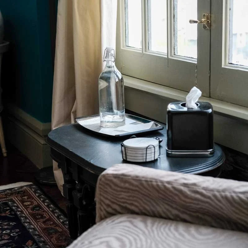 A small dark wooden table by a window holds a glass bottle of water, a metal tray, a tissue box, and a remote control, with a cozy sofa and patterned rug nearby.