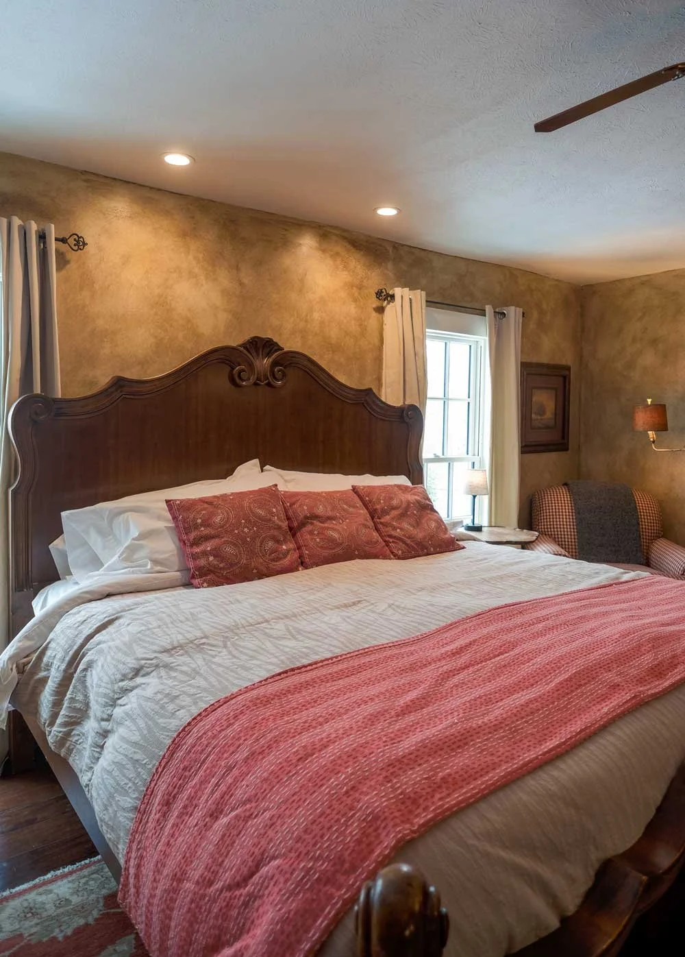 A cozy bedroom featuring a large wooden bed with a carved headboard, three red decorative pillows, a white quilt, and a pink bed runner. There is a window with white curtains, a framed picture on the wall, a small nightstand with a lamp, an armchair with a checkered fabric, and a ceiling fan.