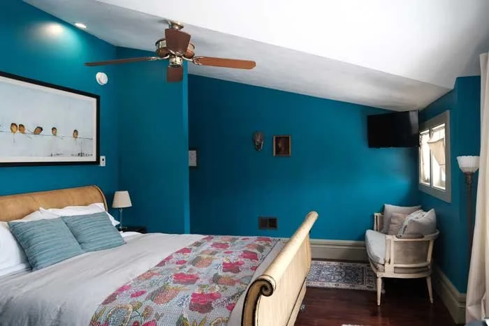 A bedroom with blue walls, a wooden bed with white and blue bedding, a small white armchair near a window, a wall-mounted TV, a ceiling fan, and wall decorations.