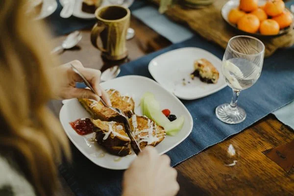 Person slicing a piece of dessert on a white plate with honey and fruit.