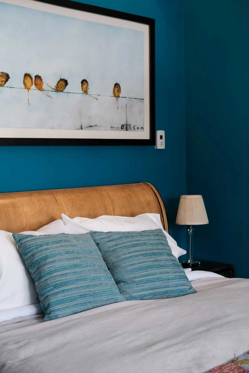A bedroom with a wooden bed frame, white bedding, and two striped teal pillows. There is a beige lamp on a side table and a large framed painting of birds on a wire hanging on a blue wall.