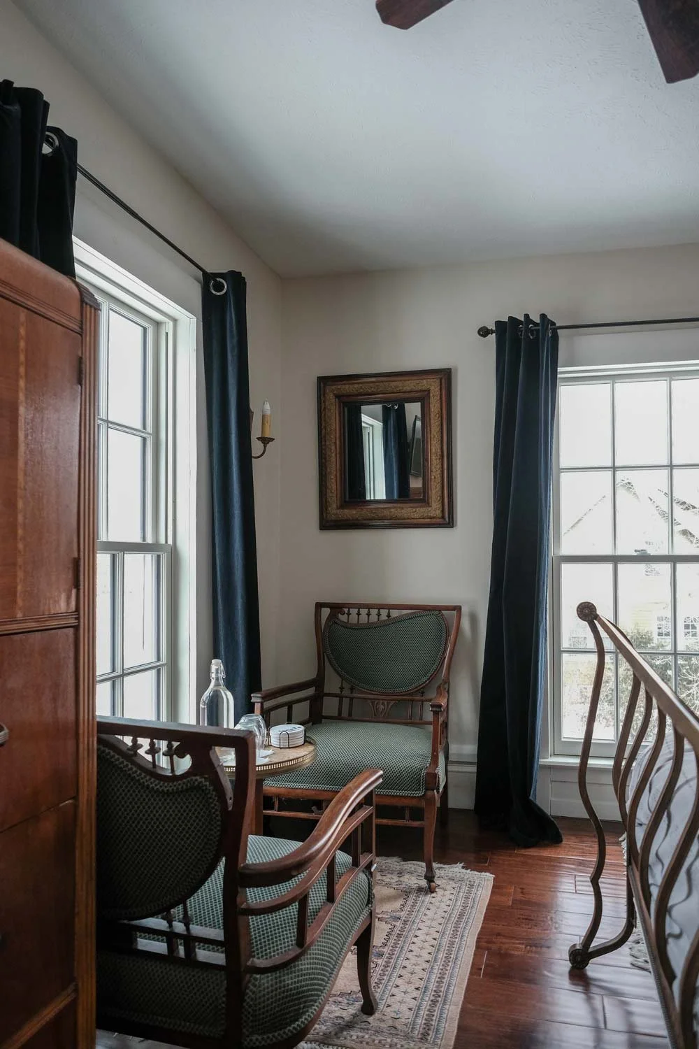 A cozy corner of a room with wooden furniture, including a chair, a small table with a water bottle, and a chest of drawers, near windows with dark curtains. There is a mirror and a patterned rug on the wooden floor.