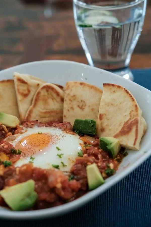 Bowl of chili topped with a poached egg and garnished with cilantro, served with sliced tortilla chips and diced avocado, with a glass of water in the background.