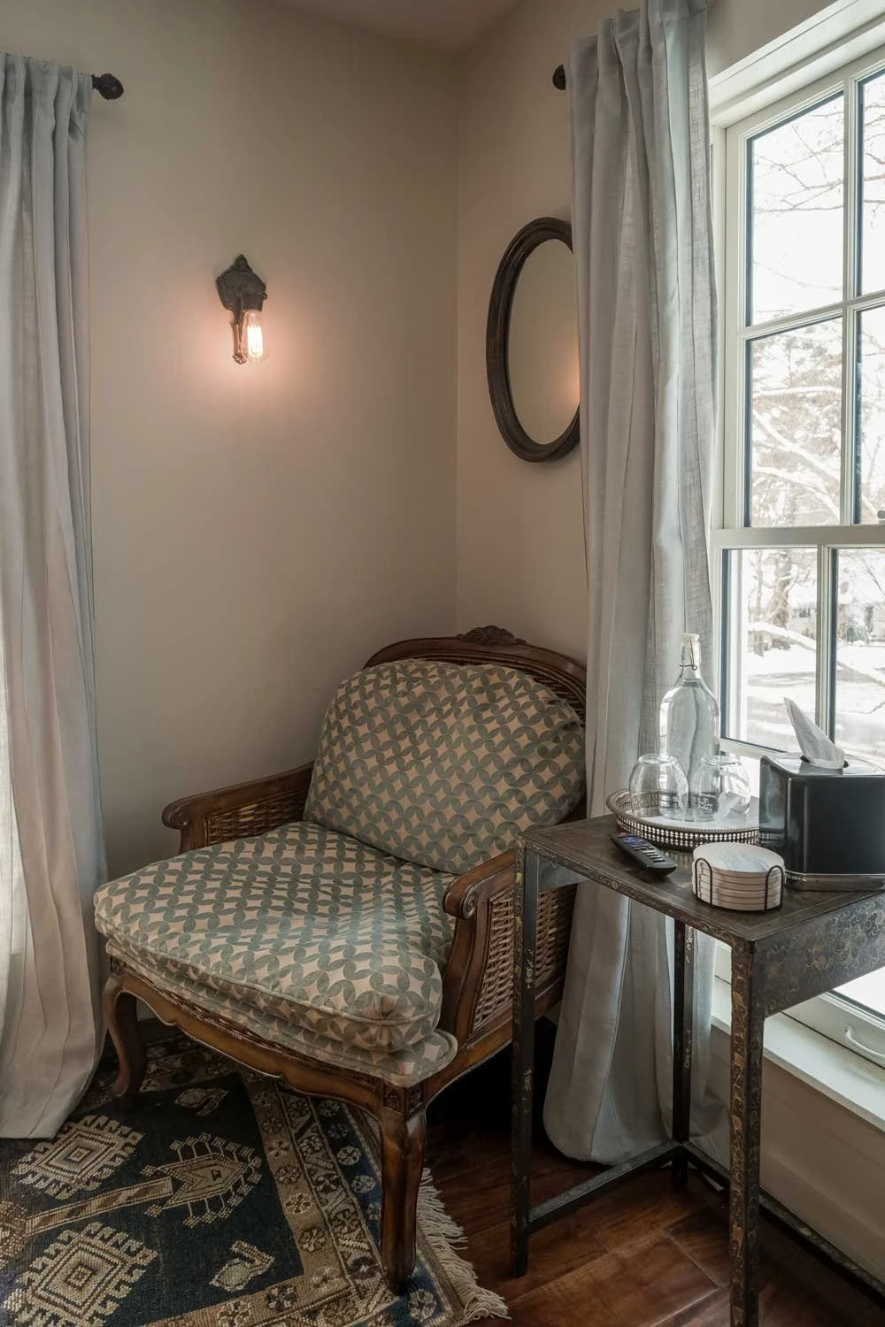 A corner of a room with a patterned armchair, a small metal side table with glasses, a remote, and tissue box, a window with white curtains, a round mirror on the wall, and an exposed light bulb