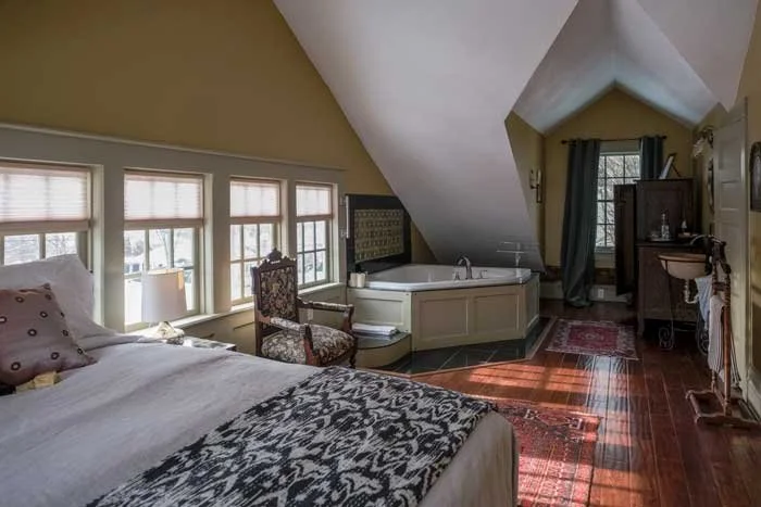 A bedroom with a sloped ceiling, multiple windows, a bed with pillows and a patterned blanket, an armchair, a corner bathtub, a dresser, and a rug on hardwood floor.