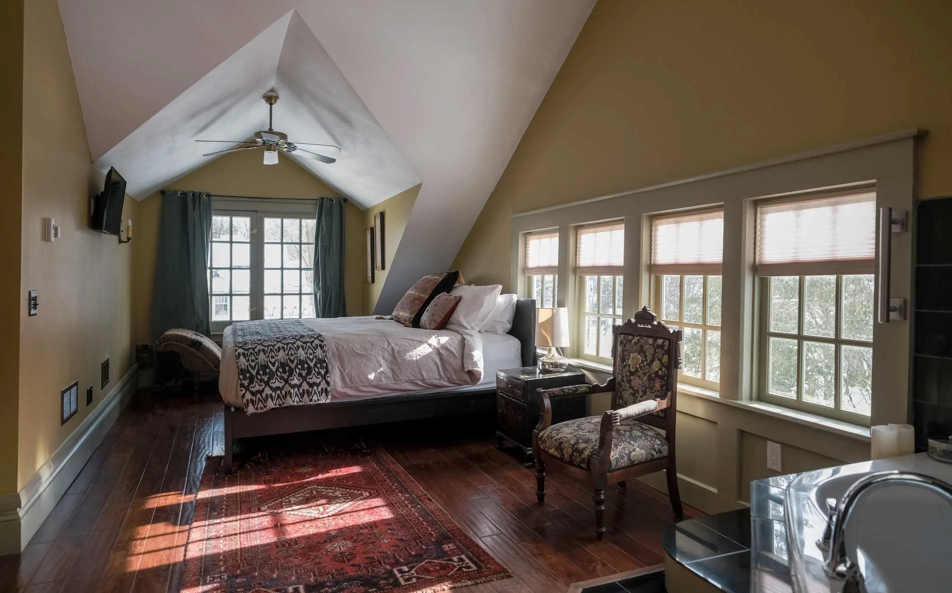 Bedroom with a sloped ceiling, a bed with pillows and a blanket, a window with curtains, and another window with blinds, a chair, a side table with a lamp, a television on the wall, a rug on the wooden floor, and sunlight coming through the windows.