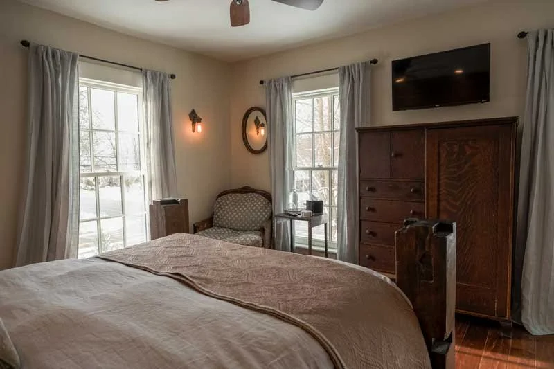 A bedroom with a bed, two windows with curtains, a wooden chest, a TV mounted on the wall, and a chair with a side table and electronic device.