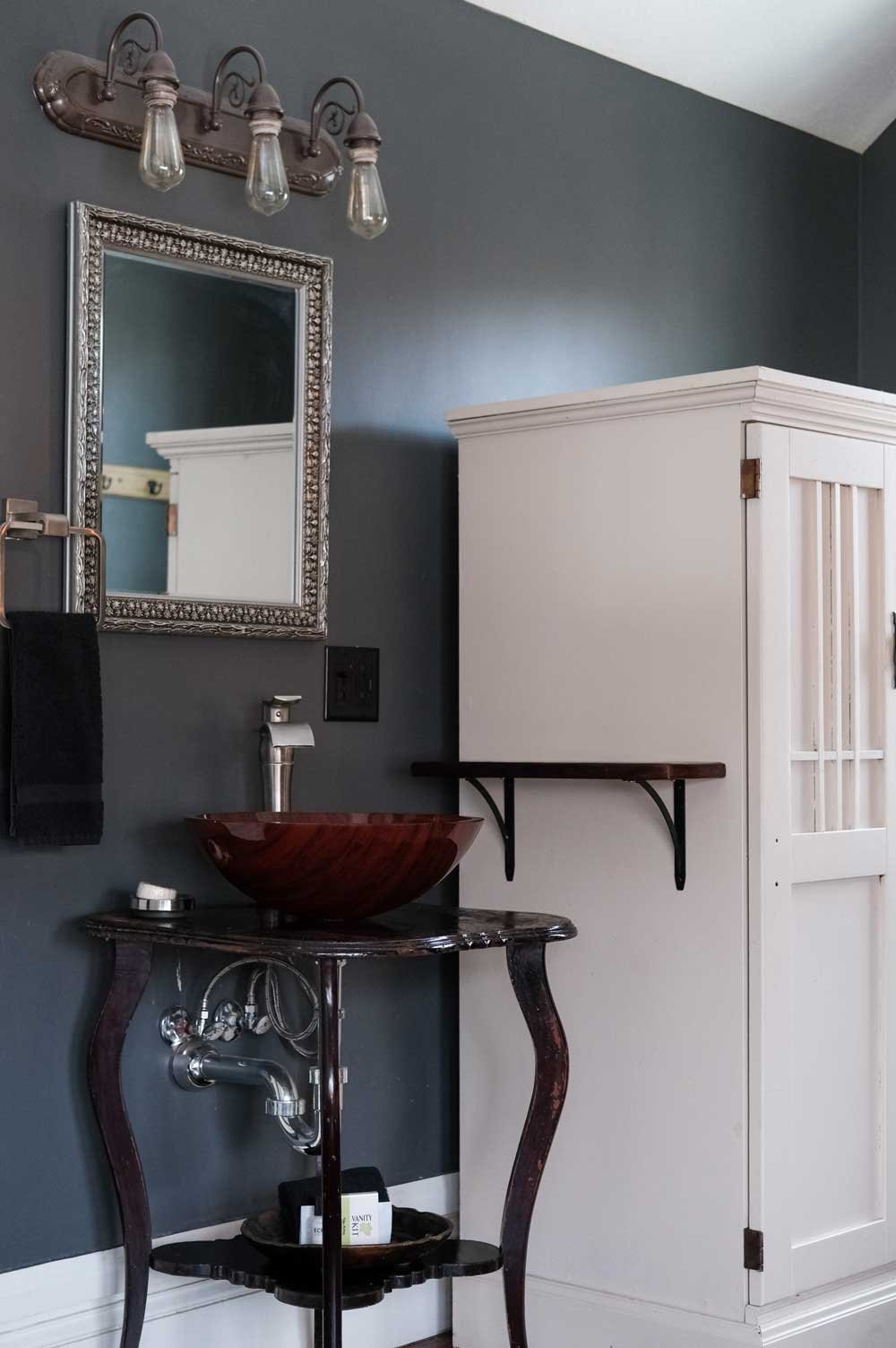 Bathroom with dark gray wall, ornate silver-framed mirror, vanity with a dark wood vessel sink, black antique-style table base, white cabinet, and bathroom fixtures.