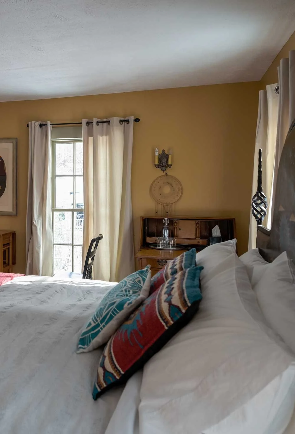 Cozy bedroom with yellow walls, white curtains, a wooden nightstand with glasses, a framed picture, and decorative pillows on a bed.