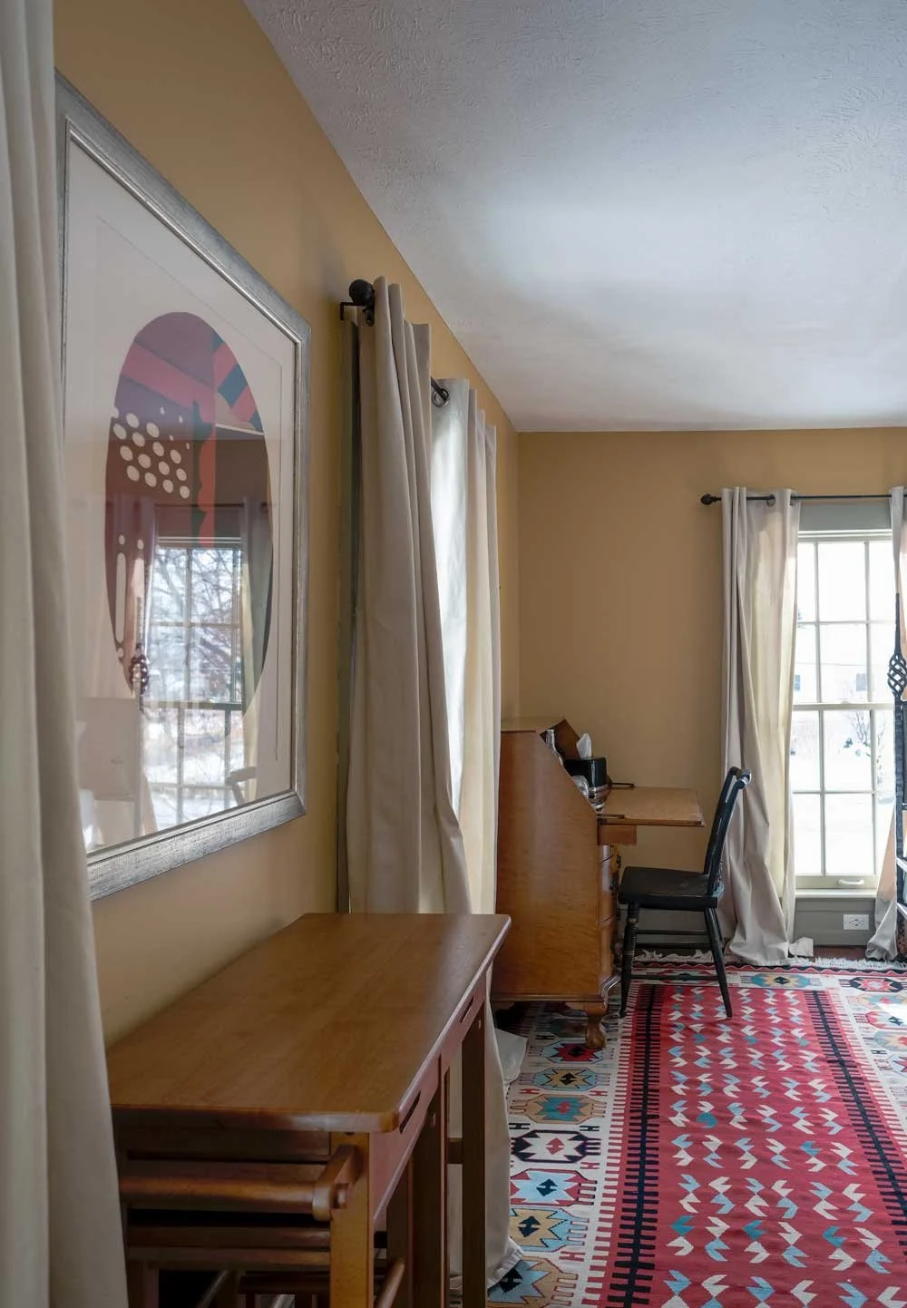 A room with beige walls and white curtains, a wooden desk, a black chair, and a colorful patterned rug, with a large window letting in natural light.