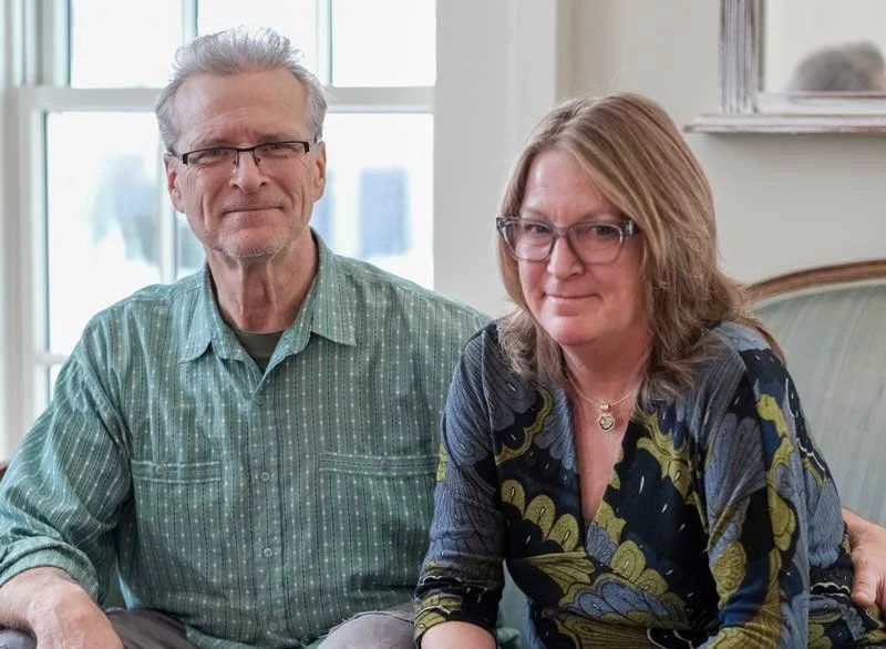 An older man and woman sitting together indoors, both smiling at the camera. The man has gray hair, glasses, and is wearing a green striped shirt. The woman has shoulder-length brown hair, glasses, and is wearing a patterned blouse with dark and yellowish colors.