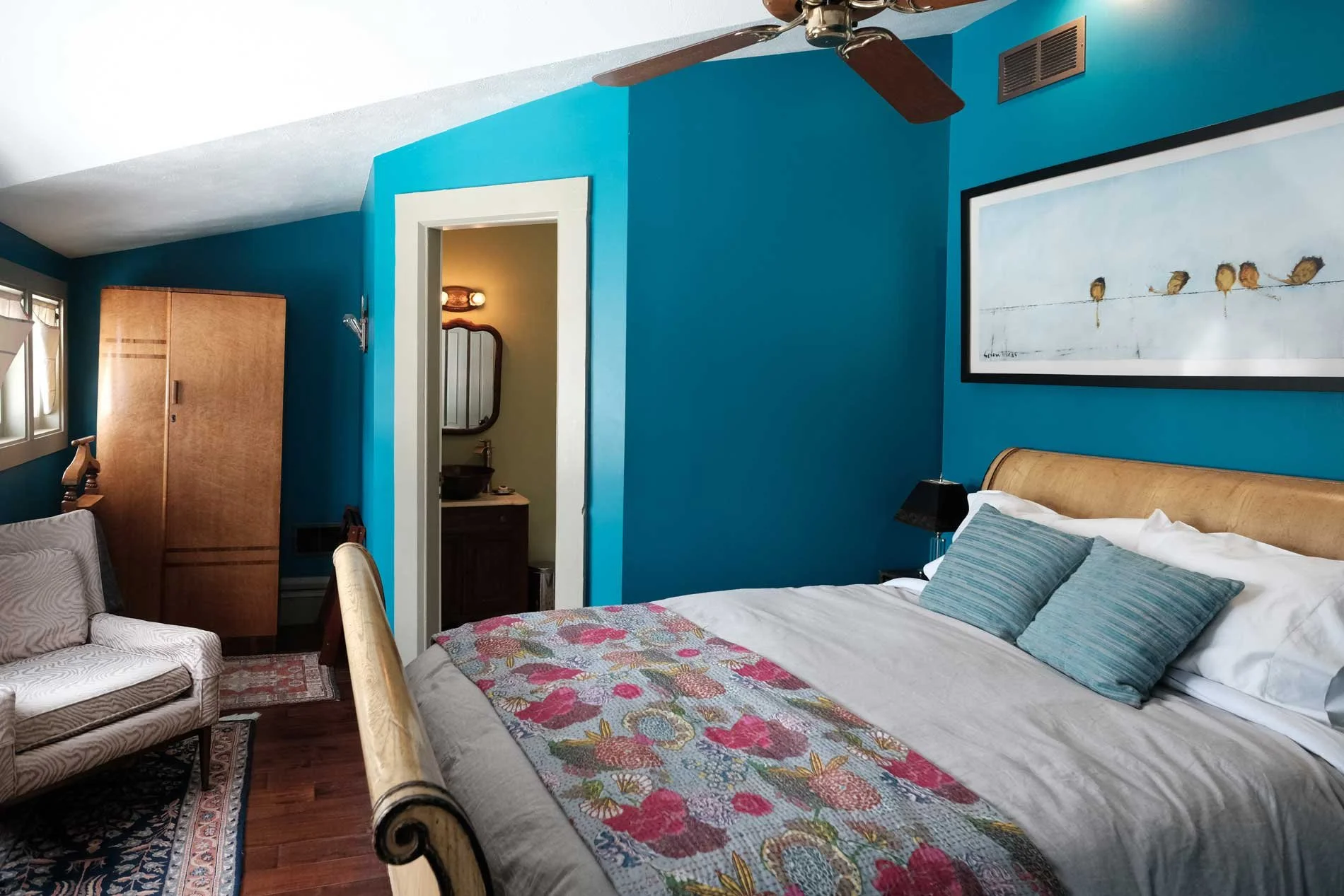 A cozy bedroom with a blue accent wall, a large bed with a wooden headboard, two teal pillows, a colorful quilt, and a black bedside lamp. There is a framed artwork of birds on a wire above the bed. A ceiling fan hangs from the ceiling. To the left, there is a window, a beige armchair, a wooden wardrobe, and a door leading to a small bathroom with a sink and mirror.