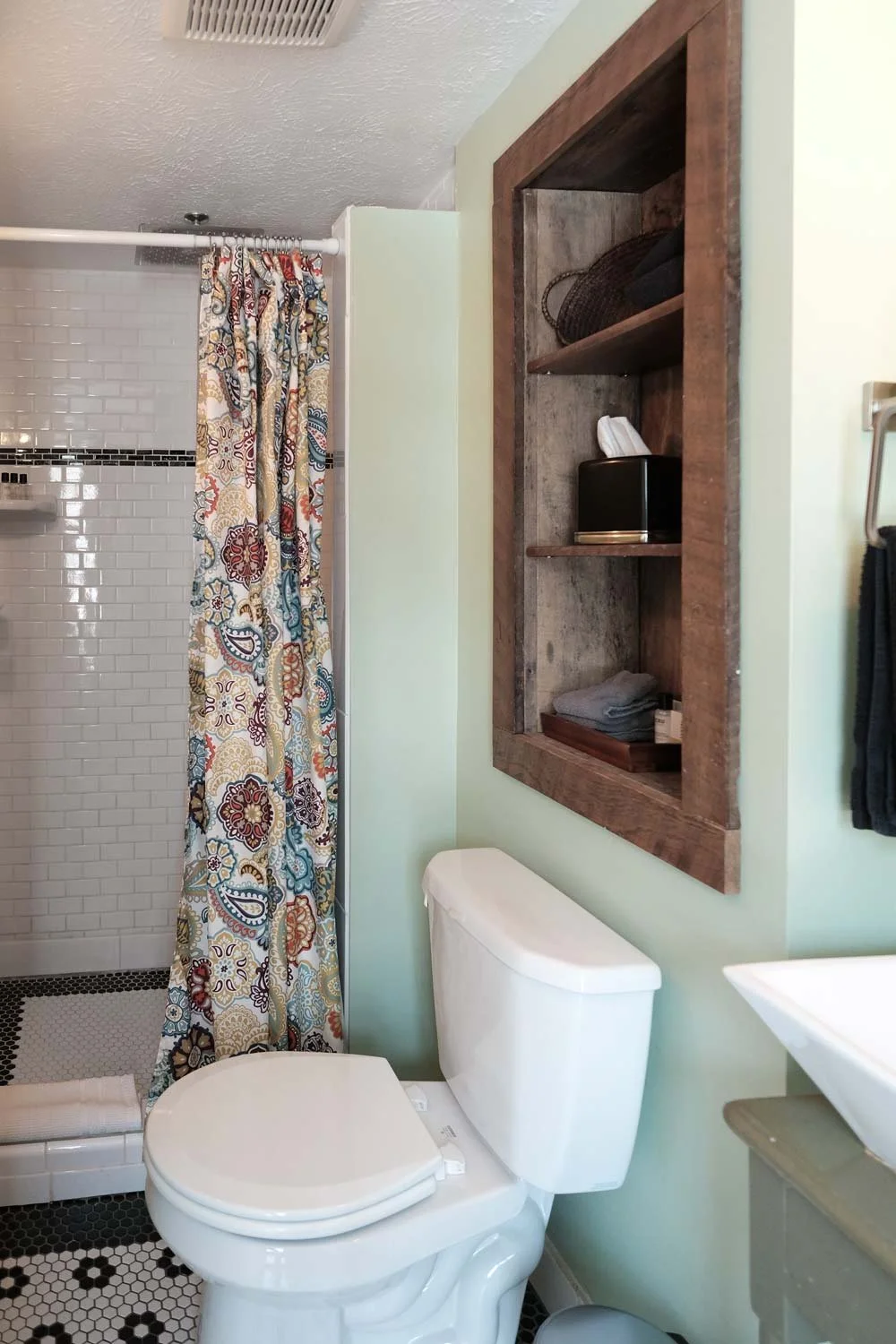 Bathroom with a white toilet, a wooden shelf cabinet on the wall with towels and a basket, patterned shower curtain, white tile shower area, and a small sink.