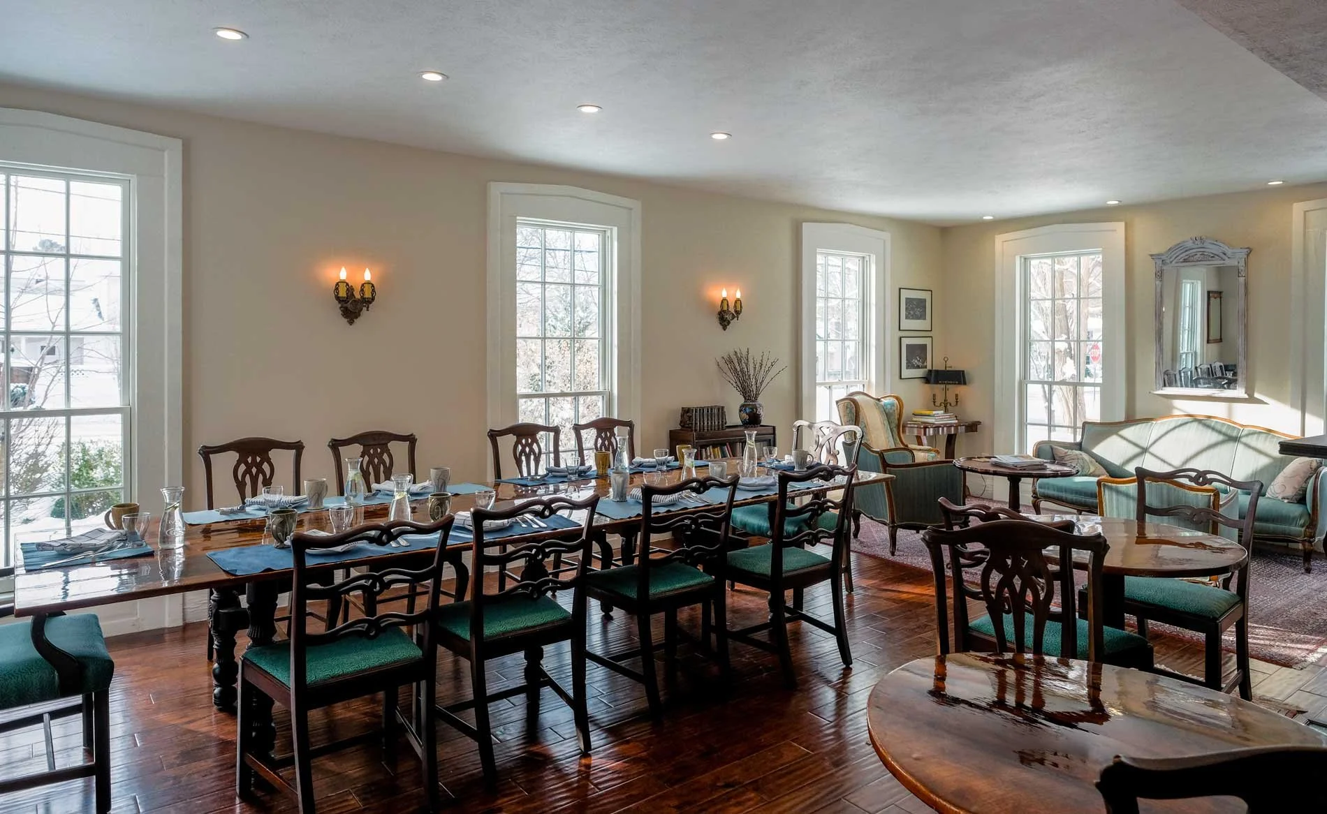 Elegantly set dining room with long wooden table, green cushioned chairs, and a cozy sitting area with antique-style furniture and large windows letting in natural light.
