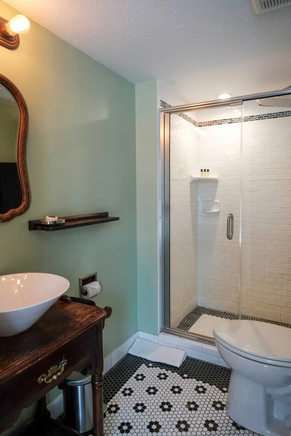 Bathroom with a glass shower, white subway tile walls, a toilet, a vintage wooden vanity with a vessel sink, a mirror, a small shelf, and patterned black and white tile flooring.