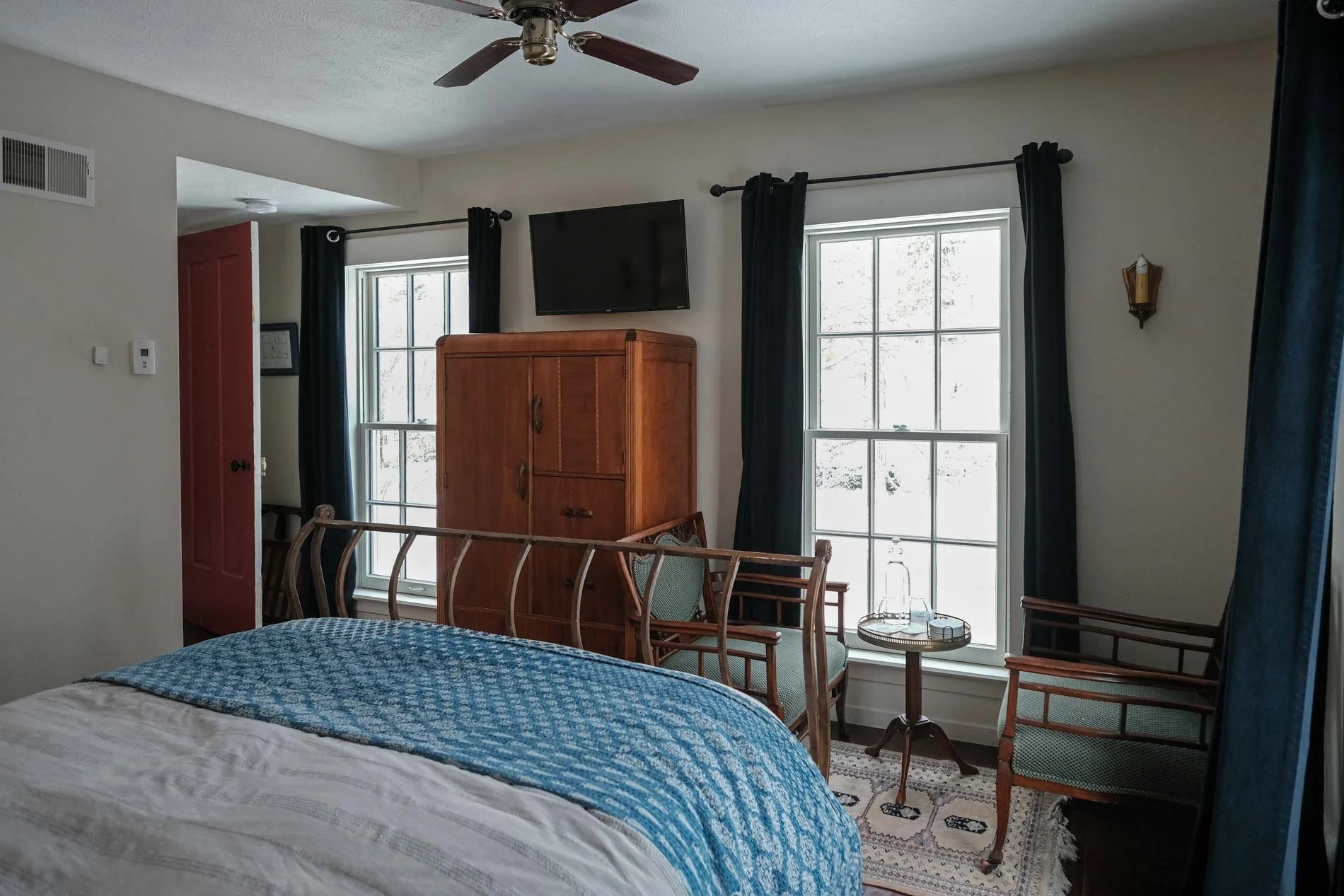 A cozy bedroom with a bed covered in a blue and white quilt, two windows with black curtains, a wall-mounted TV, an armoire, and a wooden chair beside a small round table with glassware and a tray.