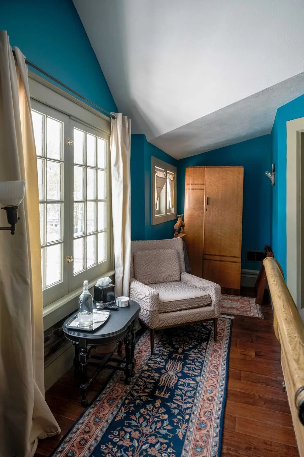 A cozy corner in a room with blue walls, a window with cream curtains, a patterned armchair, a small black side table with a bottle of water, tissue box, and remote, a wooden wardrobe, and a patterned rug on wooden floor.