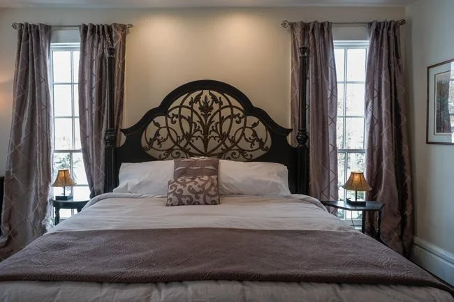 A bedroom with a decorative black wrought iron headboard, two windows with patterned curtains, nightstands with small lamps, and a framed picture on the wall.