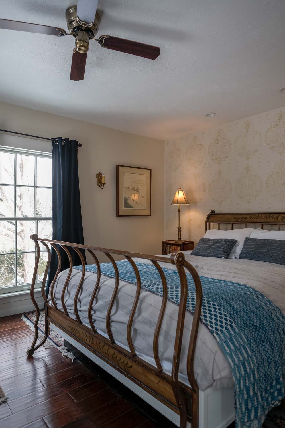 A bedroom with a wooden bed, blue and white bedding, a nightstand with a lamp, a window with dark curtains, and framed artwork on the wall.