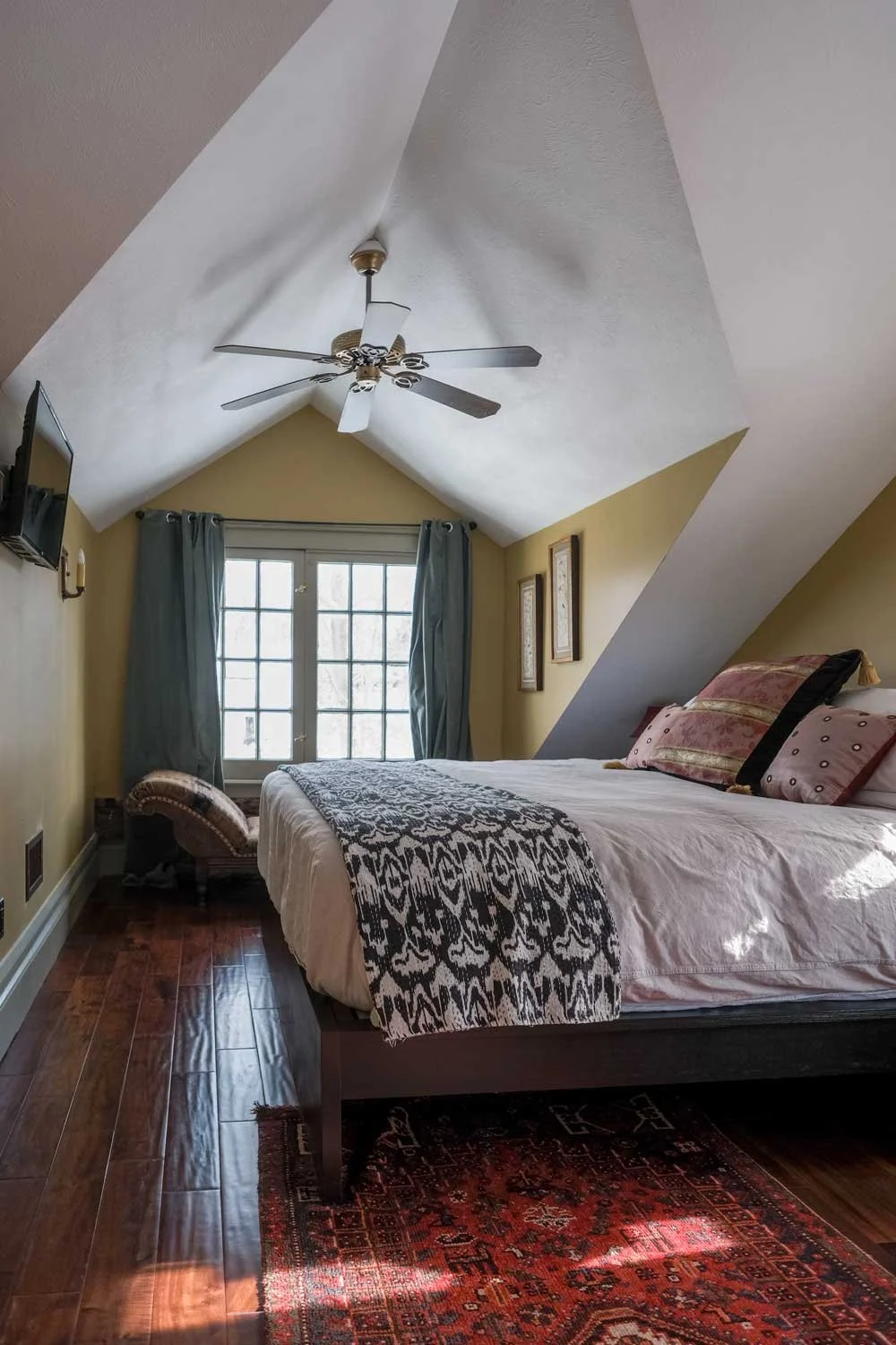 A cozy attic bedroom with a sloped ceiling, large window with blue curtains, a bed with white bedding and decorative pillows, a ceiling fan, a small bench at the foot of the bed, and framed artwork on the wall.