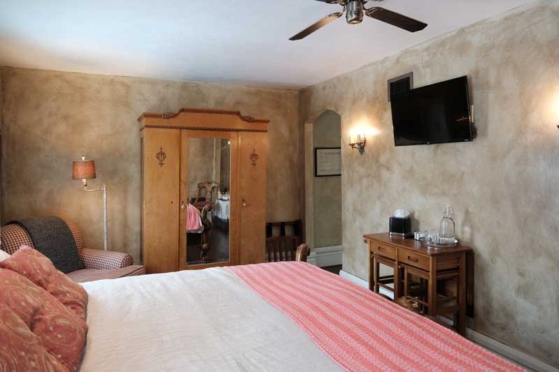 Hotel room with a bed, armchair, wooden wardrobe with mirror, wall-mounted TV, small wooden table with a water pitcher, and wall-mounted lights.