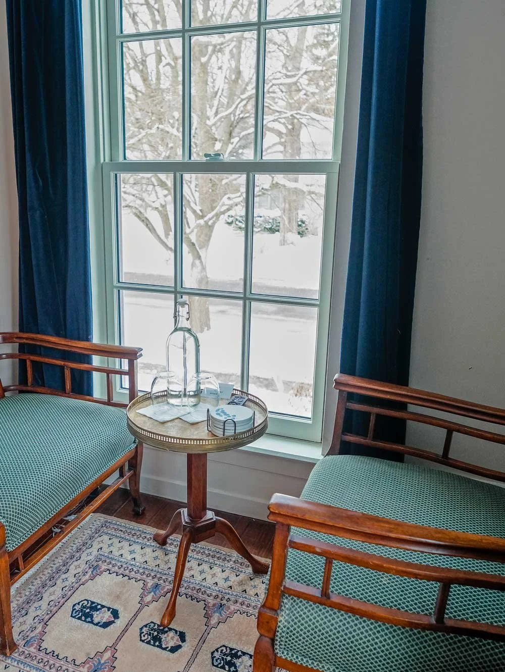 Room with a large window showing snow-covered outdoor trees, two wooden chairs with green cushions, a small round table with a glass water bottle, glasses, and coasters, and blue curtains.