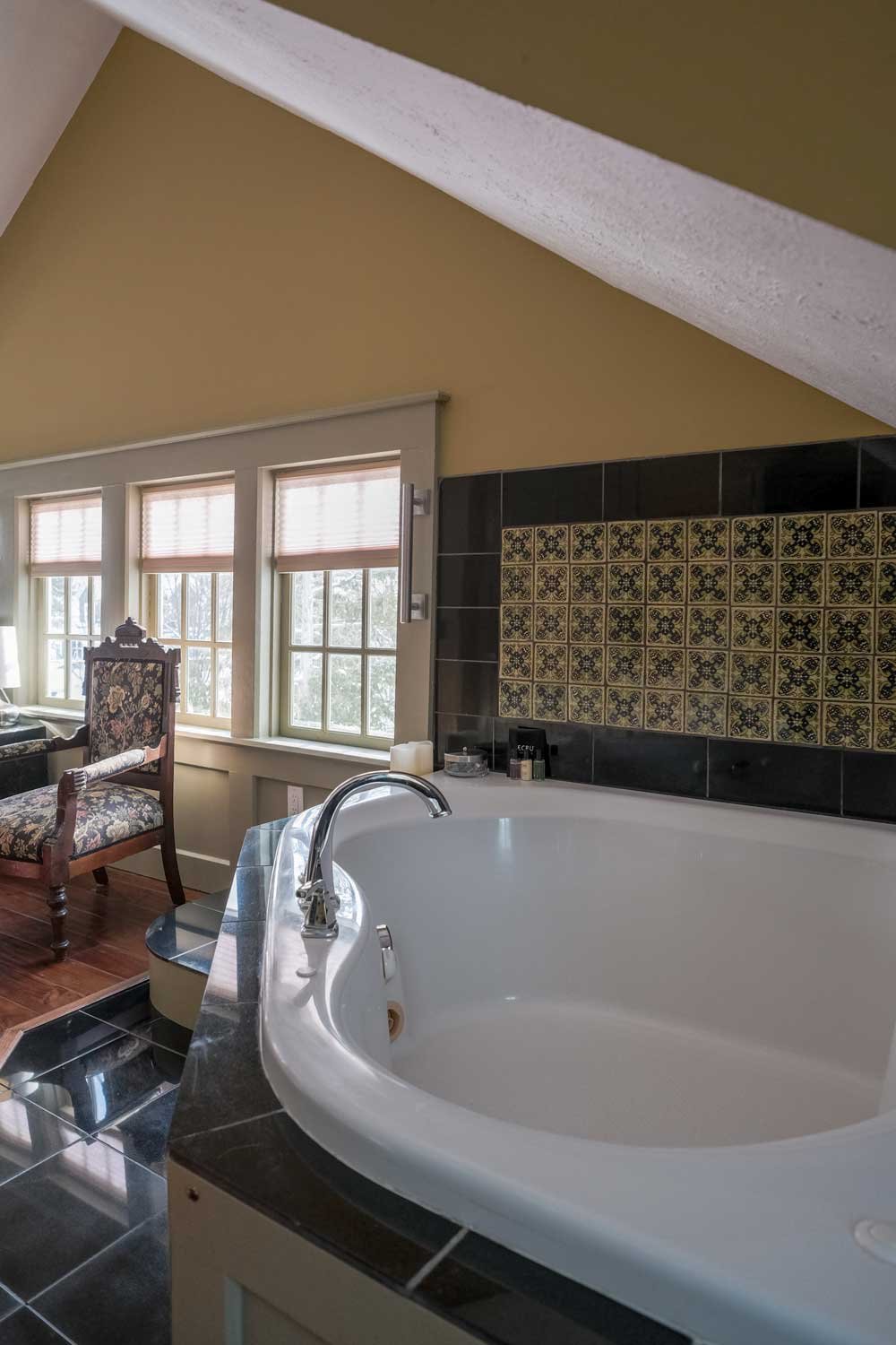 A bathroom with a white bathtub, a dark decorative tiled wall, three windows with beige blinds, and a vintage upholstered chair beside the windows.